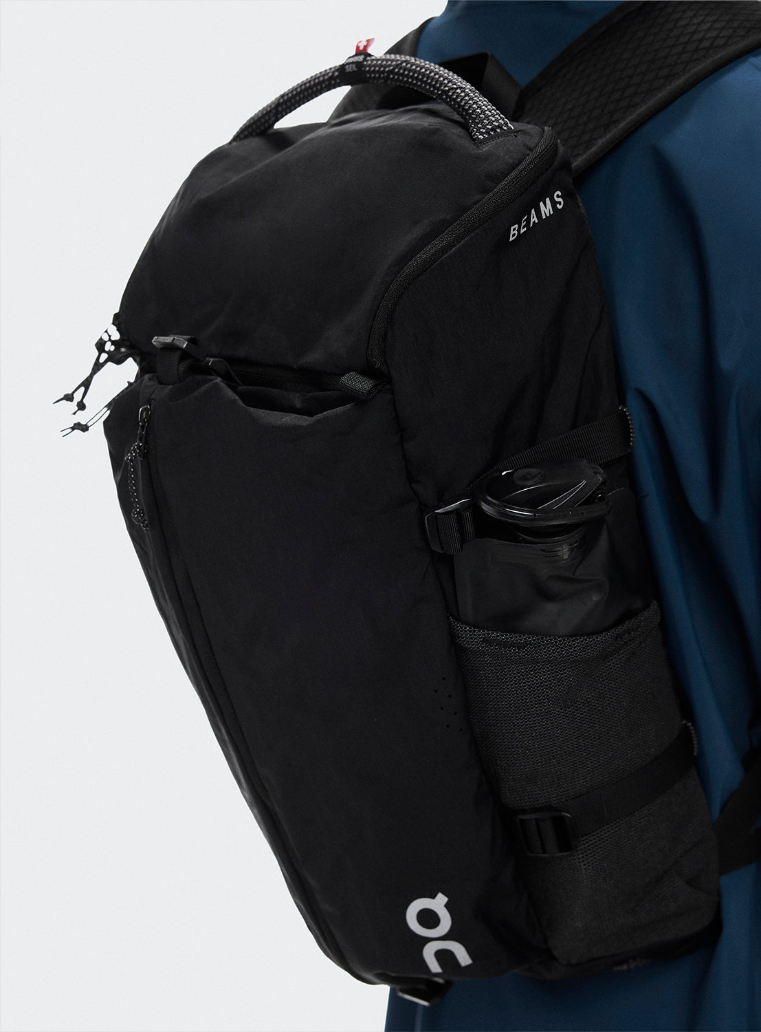 Black backpack with REI and BEAMS logo and a water bottle pocket worn by a person in a blue jacket.