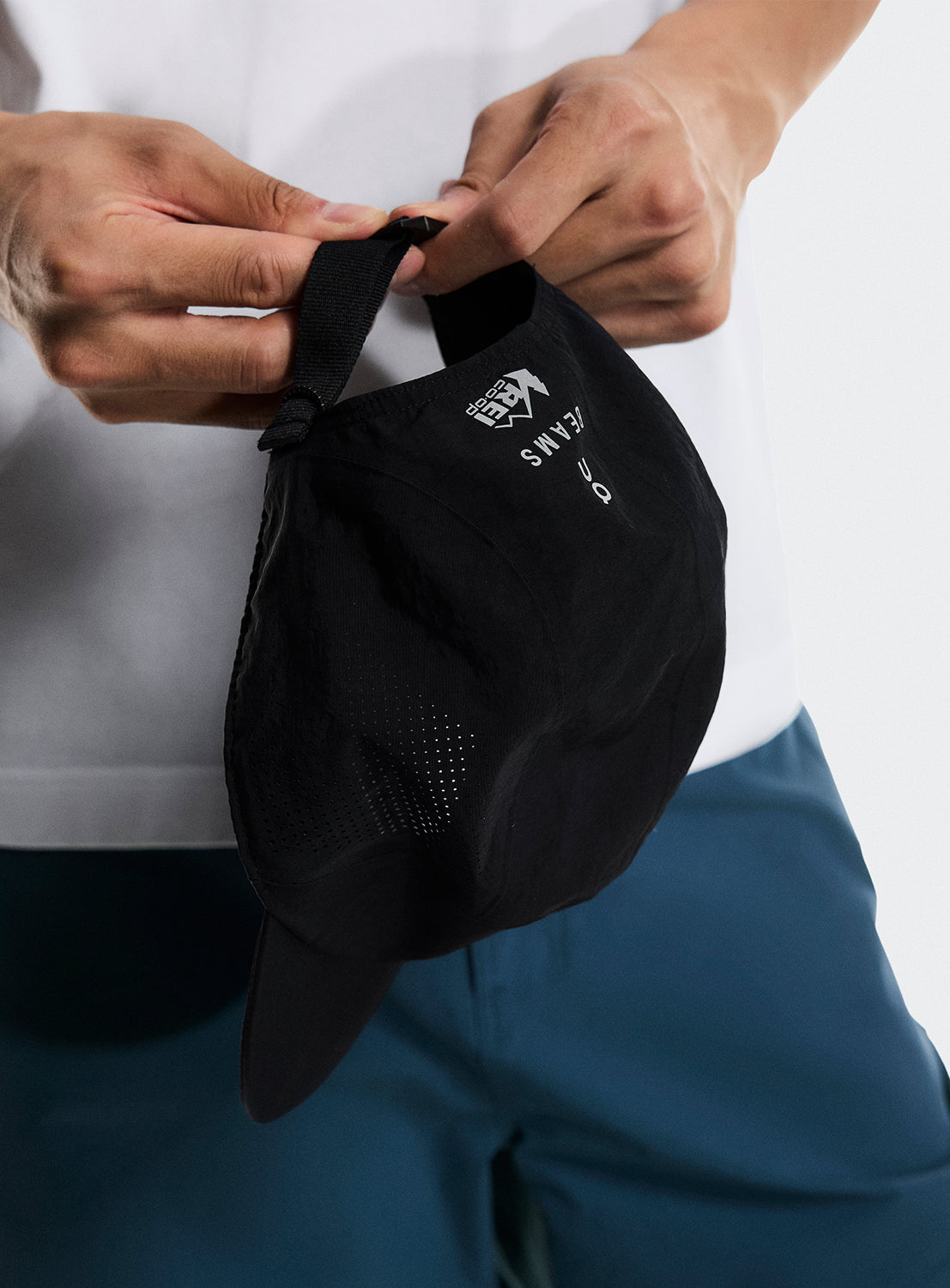 Person holding a black cap with REI CO-OP, BEAMS, AND ON Running log. 