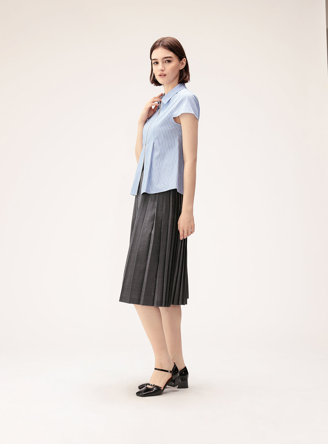 A woman stands sideways against a plain light background, wearing the BEAMS BOY Mixed Pleat Skirt in dark gray paired with a light blue short-sleeve blouse made from lightweight fabric and black heels.