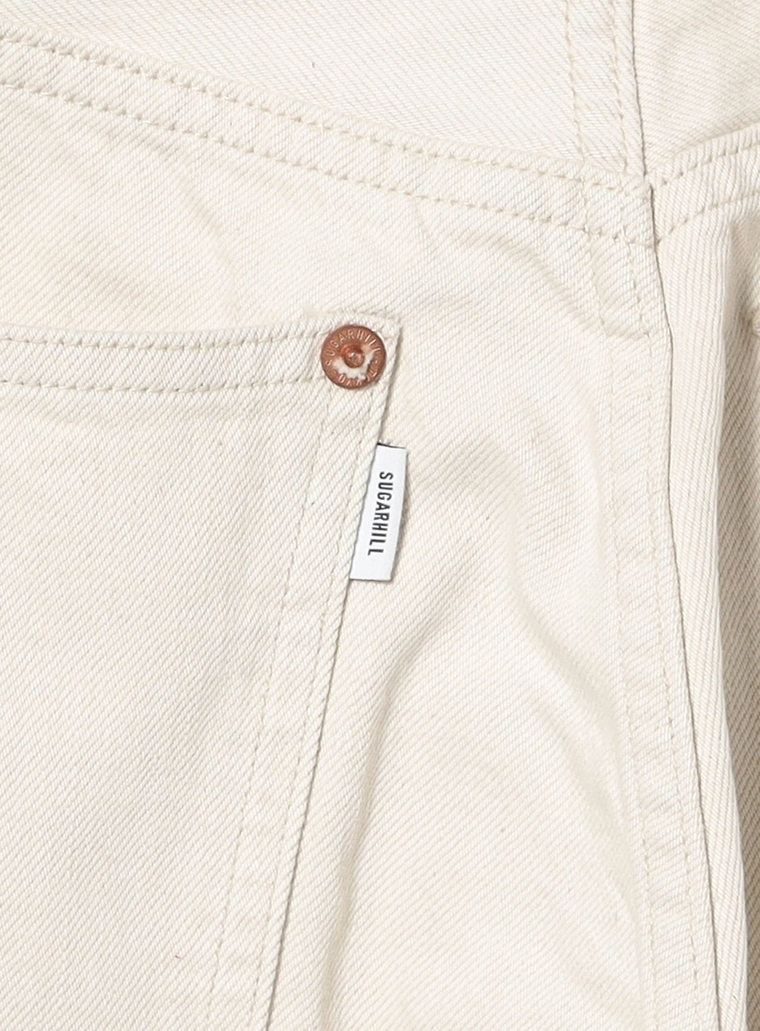 Close-up of the pocket and seam of SUGARHILL's Tattoo Flash Print Modern Denim Flared Cut jeans, cream-colored with a brown rivet and a small white SUGARHILL label.