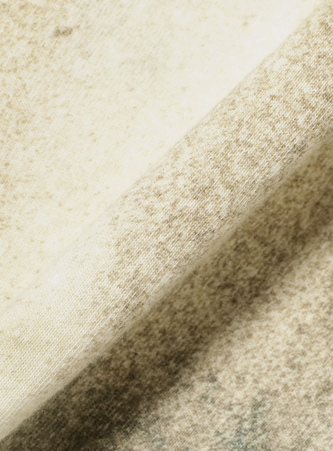 A close-up of the soft, light beige and mottled fabric of the Logo Tie Dye Tee by YUKIDYE from BAL showcases its slightly fuzzy texture, with a diagonal fold highlighting patterns reminiscent of hand-dyed YUKIDYE designs.