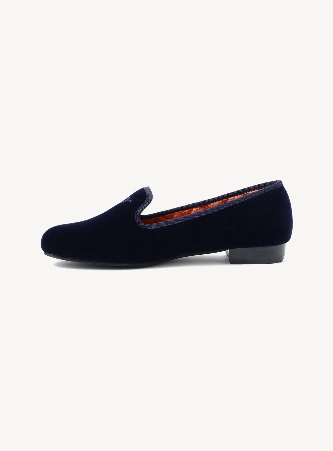 Side view of a pair of navy velvet slippers with quilted orange interior on a white background.