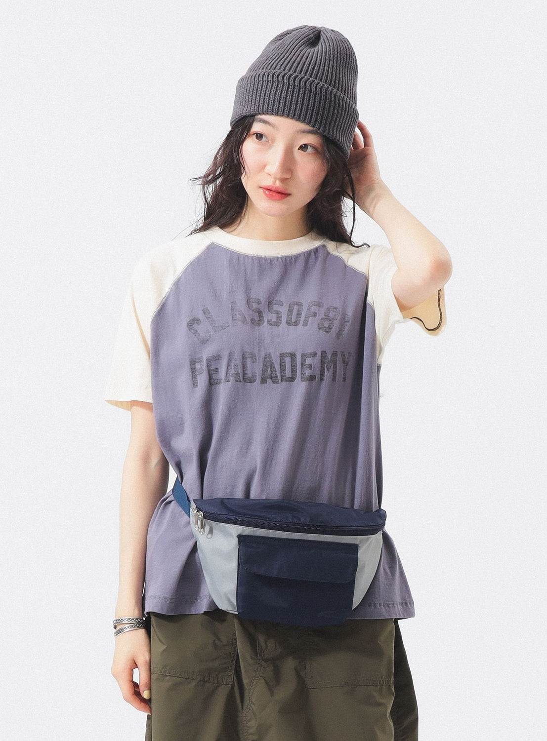 A woman in a gray beanie, olive pants, and blue-gray waist bag wears the BEAMS BOY Two-Tone Vintage Raglan T-shirt with "CLASS OF 89 PEACE ACADEMY" text, posing against a plain white background as she touches her hat and looks to the side.
