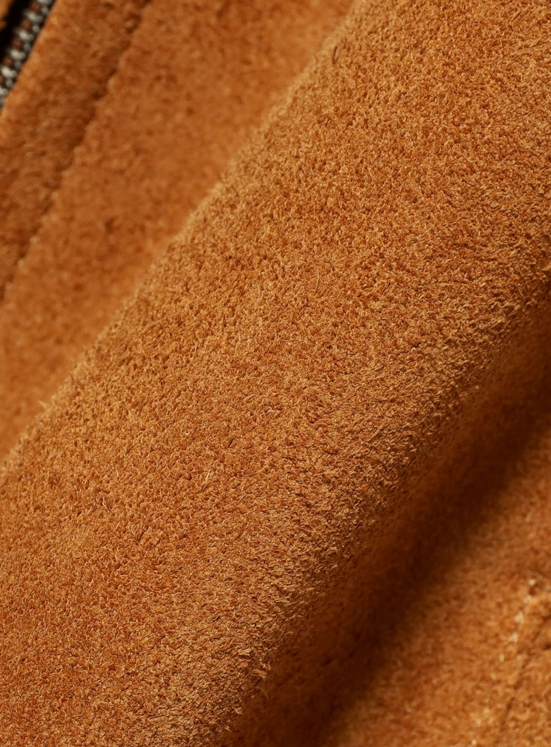 Close-up of BEAMS Suede Zip-Up Blouson in soft, textured brown suede with visible fibers and stitching. The folded material highlights its velvety surface and rich, warm color—classic details of this vintage-inspired outerwear piece.