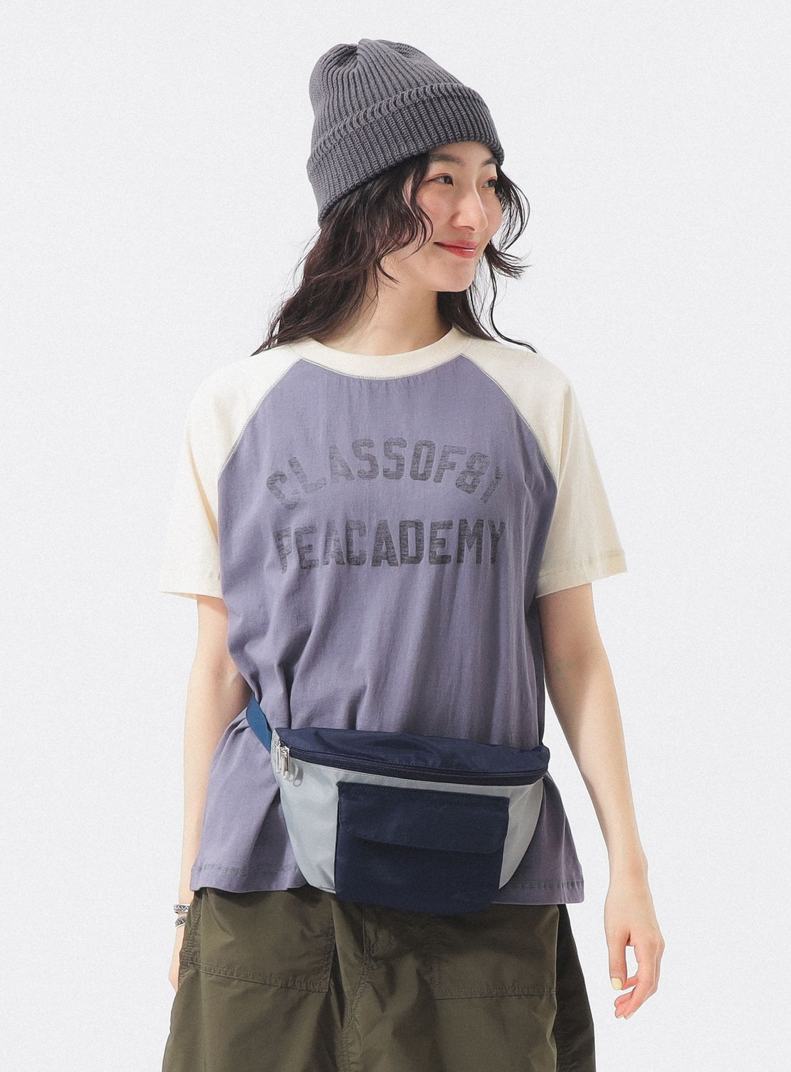 A person with long hair wears a gray beanie, green shorts, blue waist bag, and BEAMS BOY Two-Tone Vintage Raglan T-shirt, standing against a white background and smiling slightly while looking to the side.