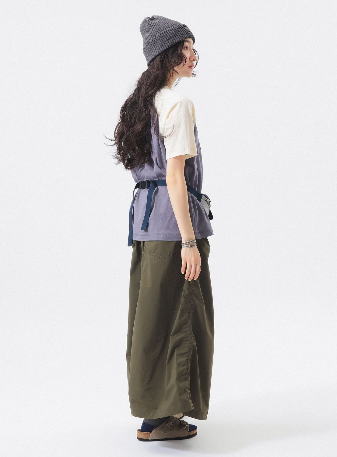 A person with long hair wears a gray beanie, BEAMS BOY Two-Tone Vintage Raglan T-shirt, blue vest, olive green wide-leg pants, and brown sandals, standing and looking over their shoulder against a plain white background.