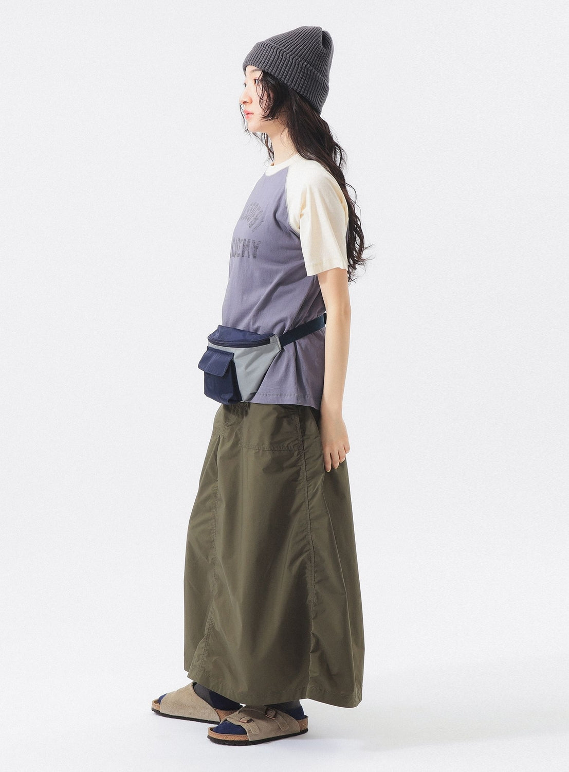 A person stands sideways in a BEAMS BOY Two-Tone Vintage Raglan T-shirt, an olive green maxi skirt, a color-blocked waist bag, and beige sandals with socks against a plain white background.