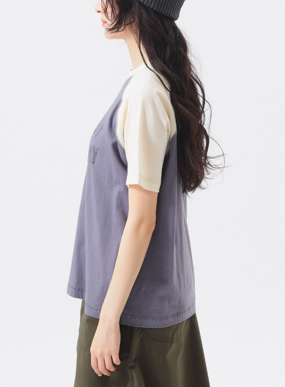 A person with long wavy hair, in a beanie and olive green pants, stands sideways against a white background wearing the BEAMS BOY Two-Tone Vintage Raglan T-shirt in light purple and cream, evoking collegiate athletic style.