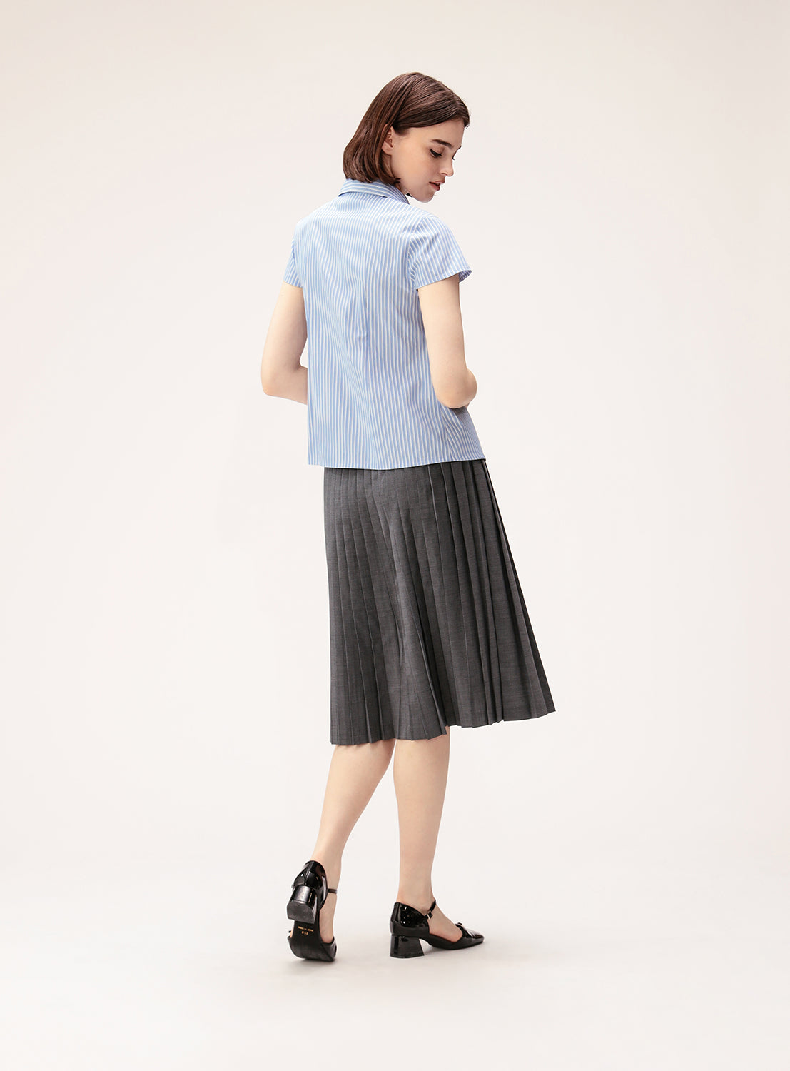A woman stands facing away, wearing a short-sleeved, light blue striped shirt paired with the BEAMS BOY Mixed Pleat Skirt—a gray pleated skirt crafted from lightweight fabric—and black shoes, set against a plain light background to highlight the ensemble’s versatile styling.
