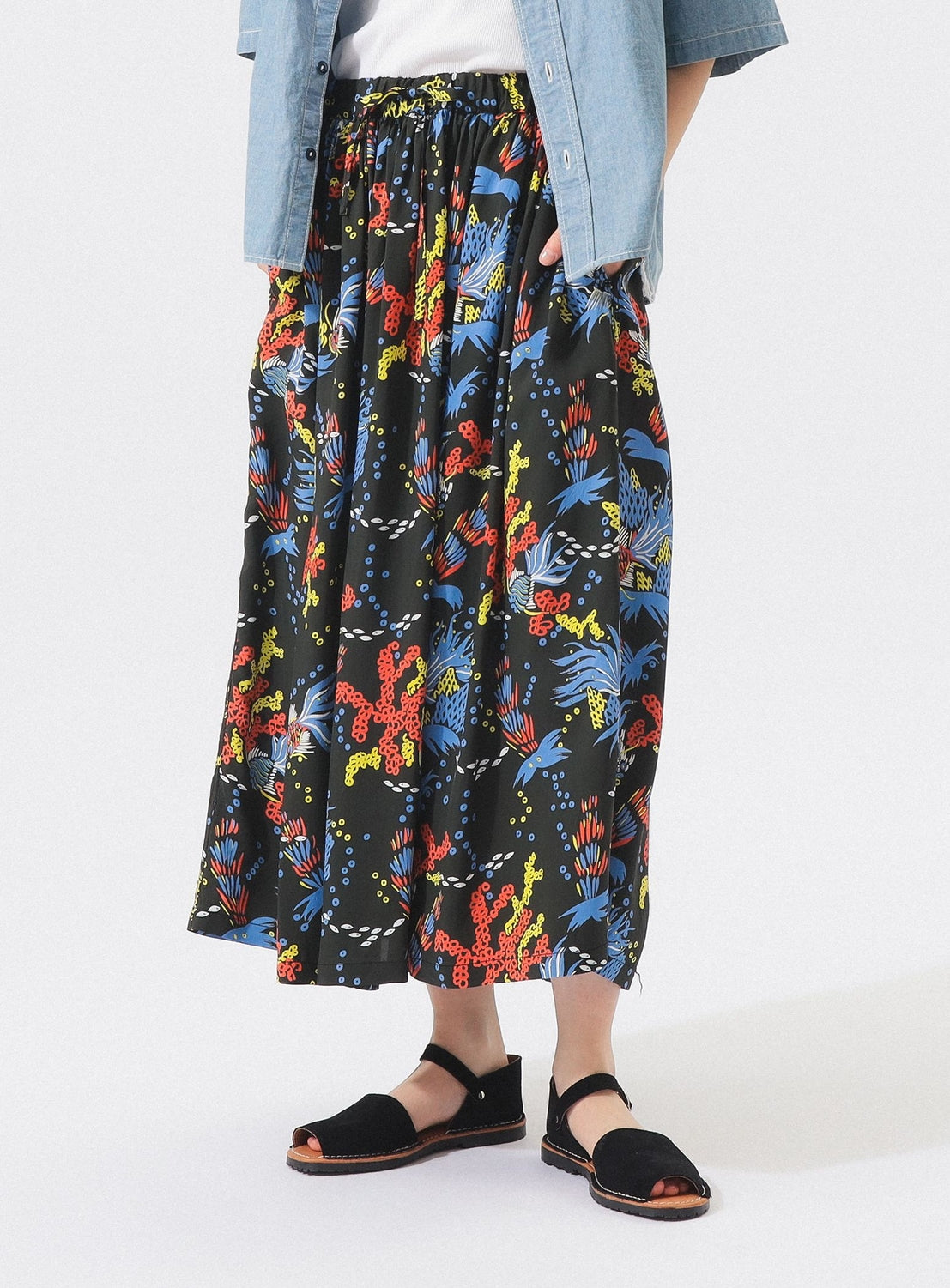 A person in a light blue shirt and black open-toe sandals stands against a plain background, hands in pockets of the [Exclusive] SUN SURF x BEAMS BOY Aloha Gathered Long Skirt from SUN SURF, featuring vintage Hawaiian prints.