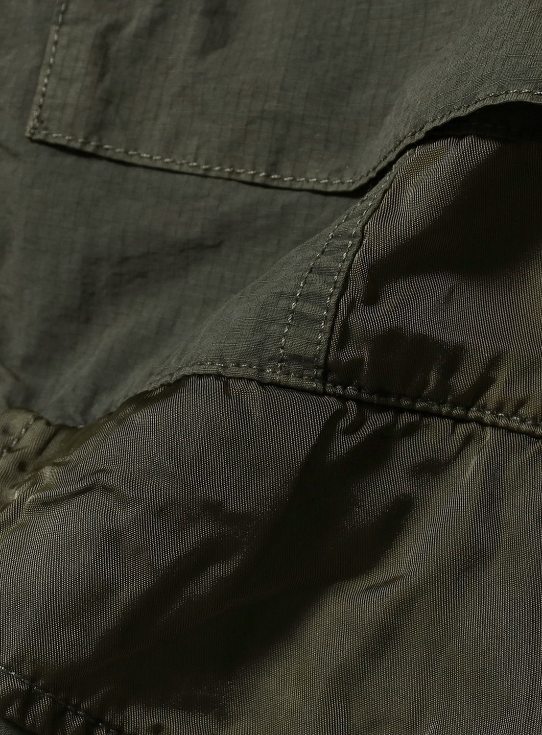 Close-up of the BEAMS Multi-Fabric Garment-Dyed M-65 Jacket, highlighting inner lining, outer pocket stitching, and detailed seams with shadows across its fabric.
