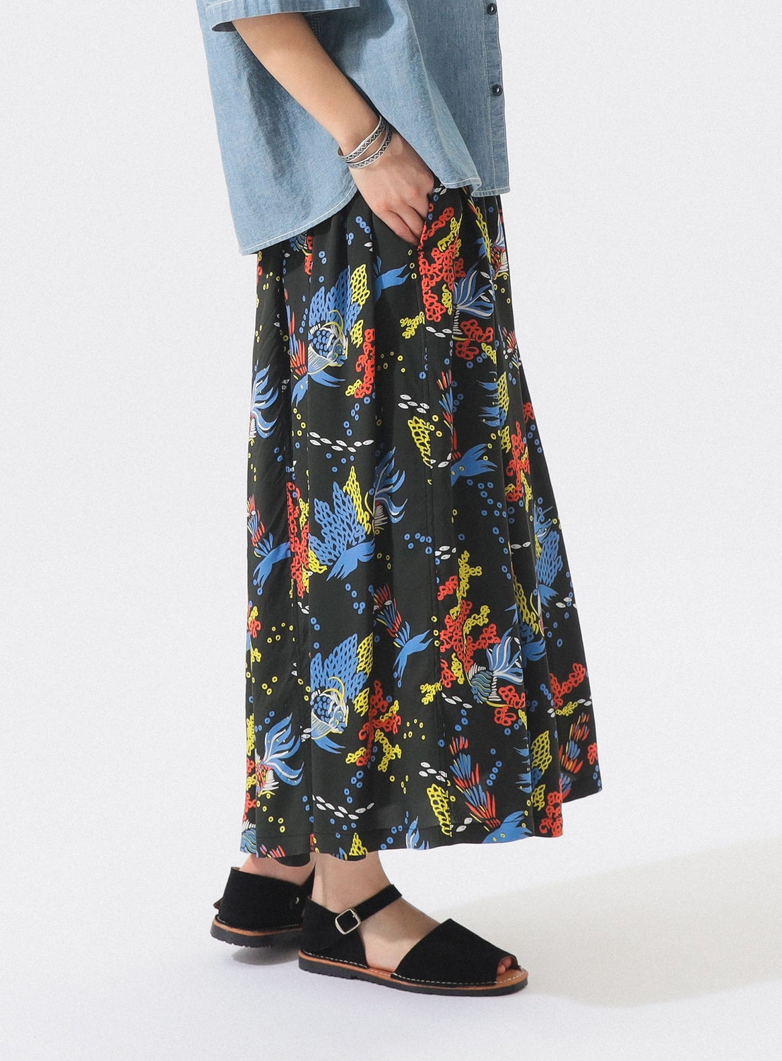A person stands against a plain white background wearing a light blue short-sleeve shirt and the [Exclusive] SUN SURF x BEAMS BOY Aloha Gathered Long Skirt by SUN SURF, paired with black open-toe ankle-strap sandals.