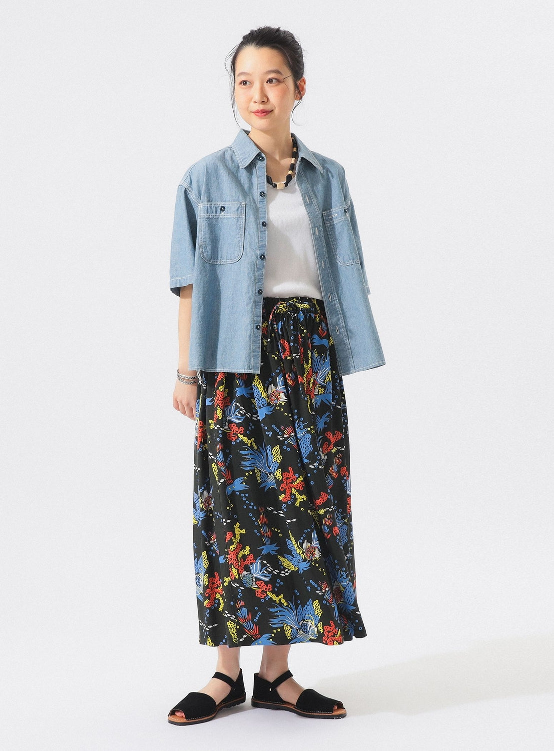 A person stands against a white background wearing a light blue denim shirt, white top, black sandals, chunky necklace, and the [Exclusive] SUN SURF x BEAMS BOY Aloha Gathered Long Skirt by SUN SURF with vintage Hawaiian prints.