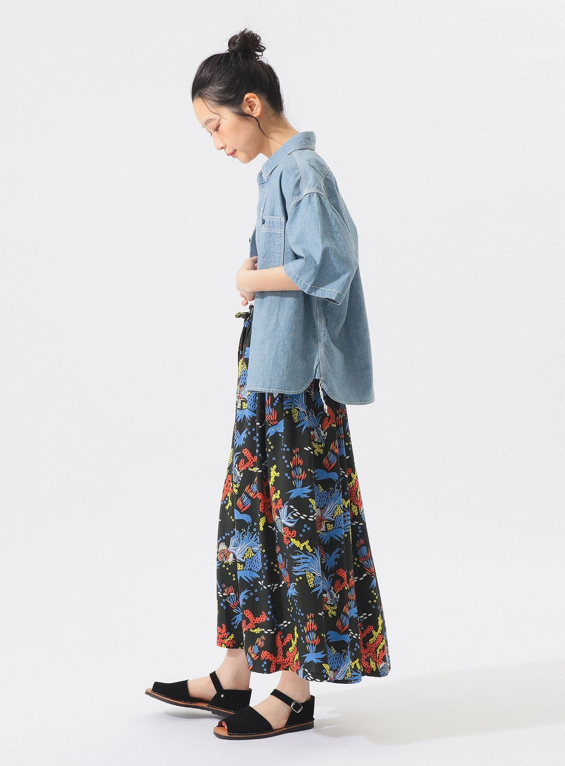 A person stands in profile against a white background, wearing a light blue short-sleeve shirt, black sandals, and the [Exclusive] SUN SURF x BEAMS BOY Aloha Gathered Long Skirt by SUN SURF. Their hair is tied up in a bun.