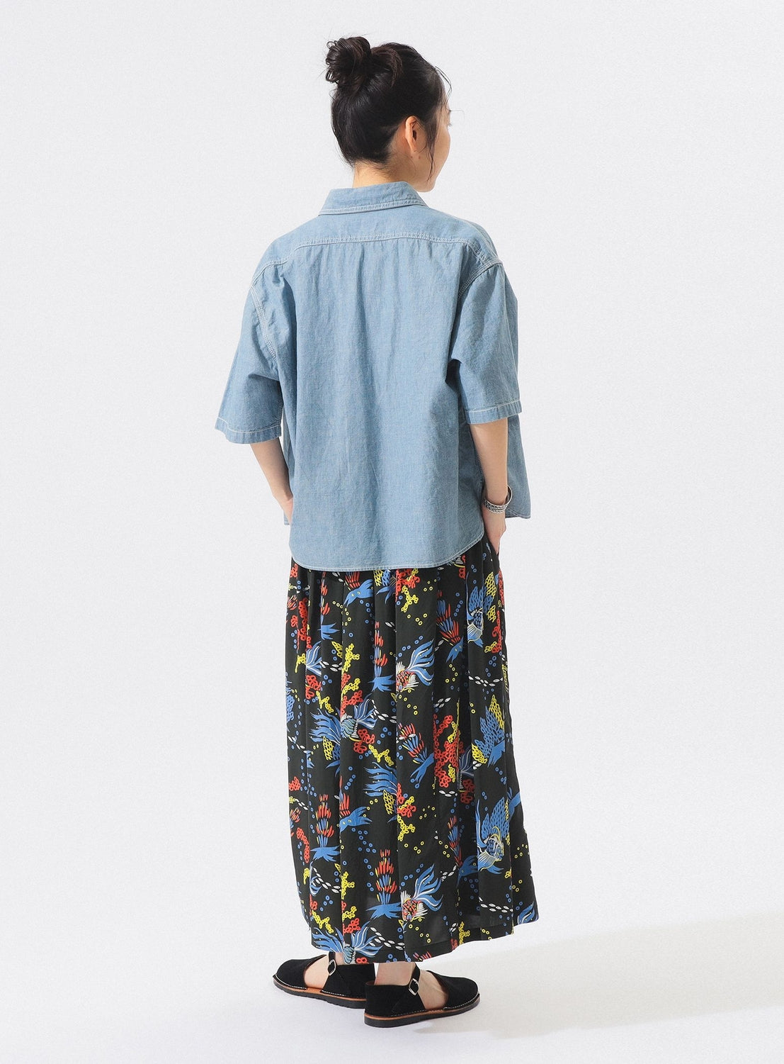 A person with hair in a bun wears a light denim short-sleeve shirt and the [Exclusive] SUN SURF x BEAMS BOY Aloha Gathered Long Skirt by SUN SURF, featuring colorful vintage Hawaiian prints. Paired with black sandals, against a plain white background.