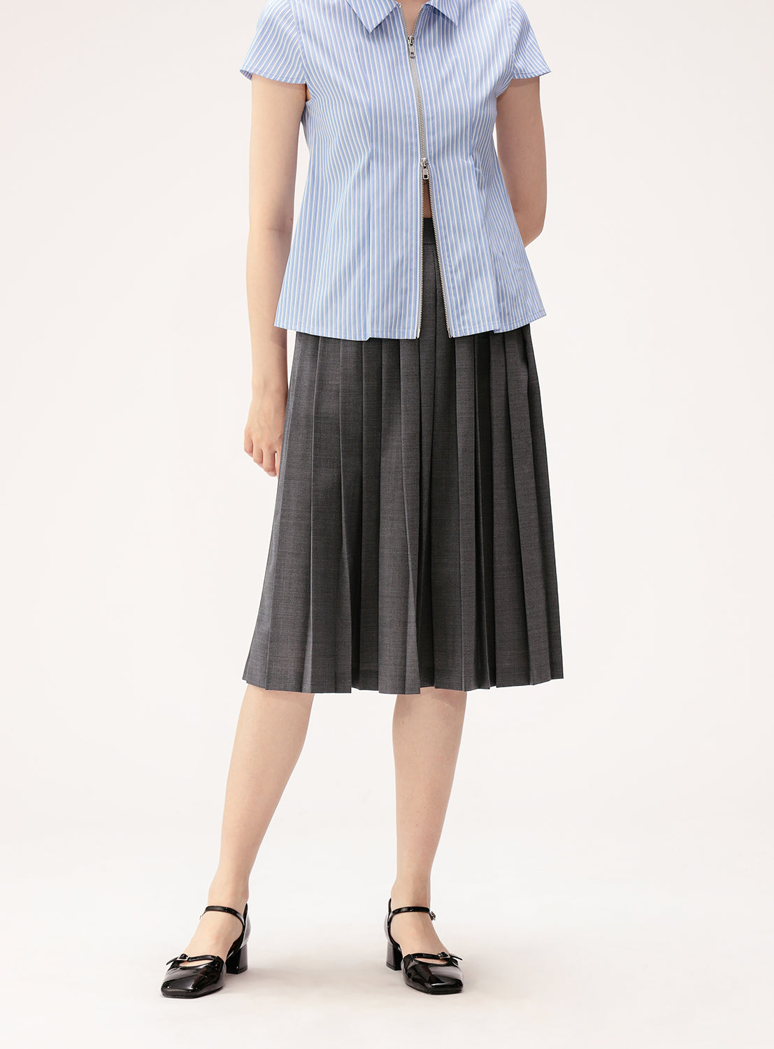 A person, with their face not visible, stands against a plain background wearing a short-sleeved, light blue pinstriped blouse paired with the BEAMS BOY Mixed Pleat Skirt—a knee-length, dark grey pleated skirt crafted from lightweight fabric—and black Mary Jane shoes.