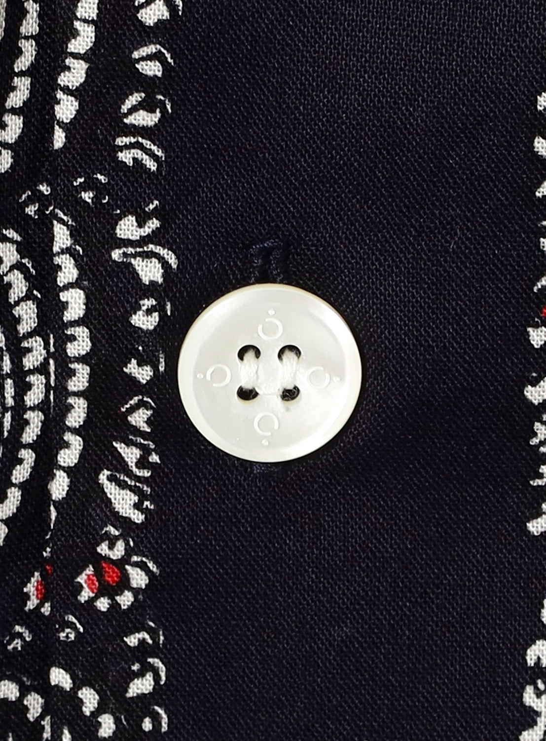 A close-up of a white button sewn onto dark fabric featuring a black, white, and red paisley print highlights the intricate details characteristic of the NOMARHYTHM TEXTILE Paisley Open Collar Shirt.