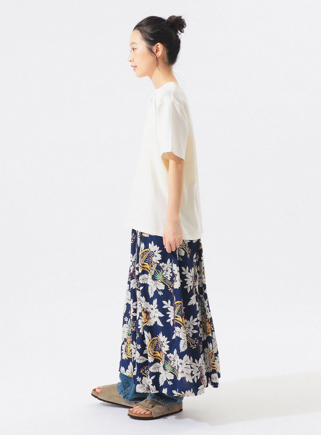 A woman stands in profile against a white background, wearing a white short-sleeve top, the [Exclusive] SUN SURF x BEAMS BOY Aloha Gathered Long Skirt with vintage Hawaiian prints, and beige sandals. Her hair is tied in a bun.