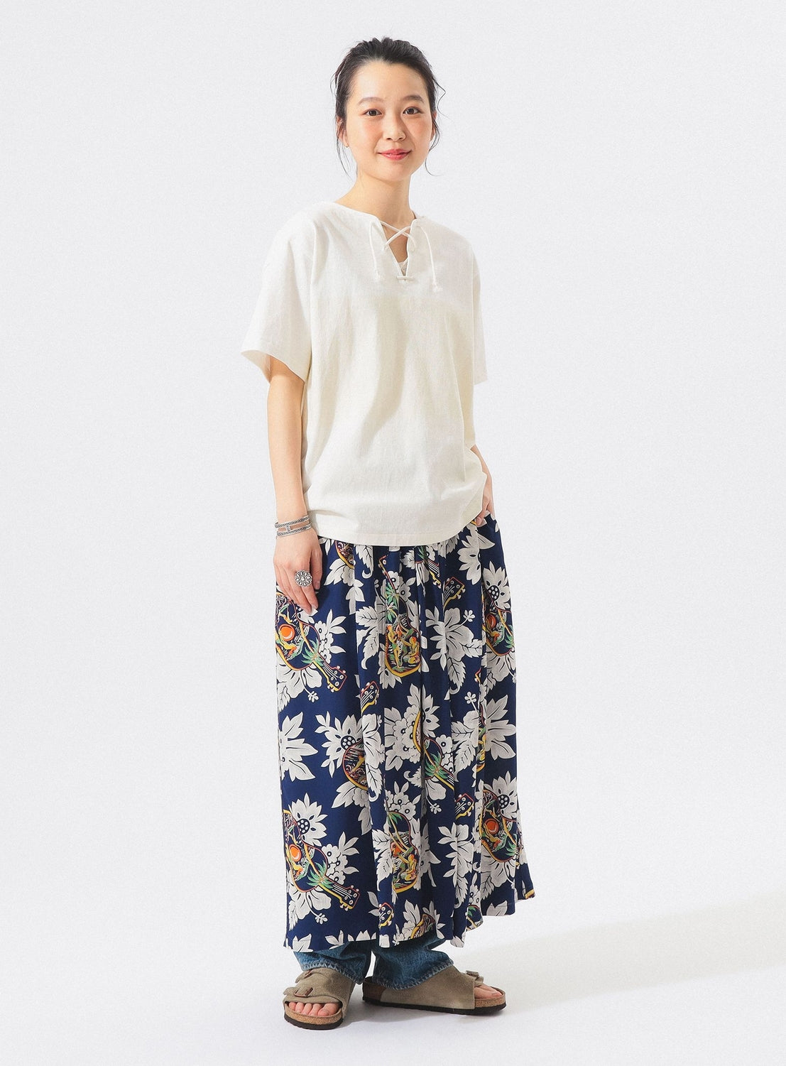 A person stands smiling in a loose white short-sleeve top, layered jeans, gray sandals, and the [Exclusive] SUN SURF x BEAMS BOY Aloha Gathered Long Skirt by SUN SURF, featuring vintage-inspired Hawaiian prints, against a plain white background.