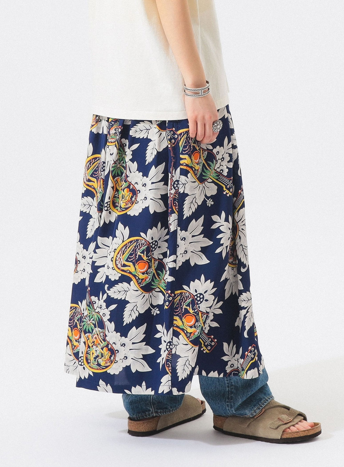 A person wears a white shirt, blue jeans, tan sandals, silver bracelets, and the [Exclusive] SUN SURF x BEAMS BOY Aloha Gathered Long Skirt by SUN SURF. The image is cropped at the shoulders to highlight this stylish outfit.