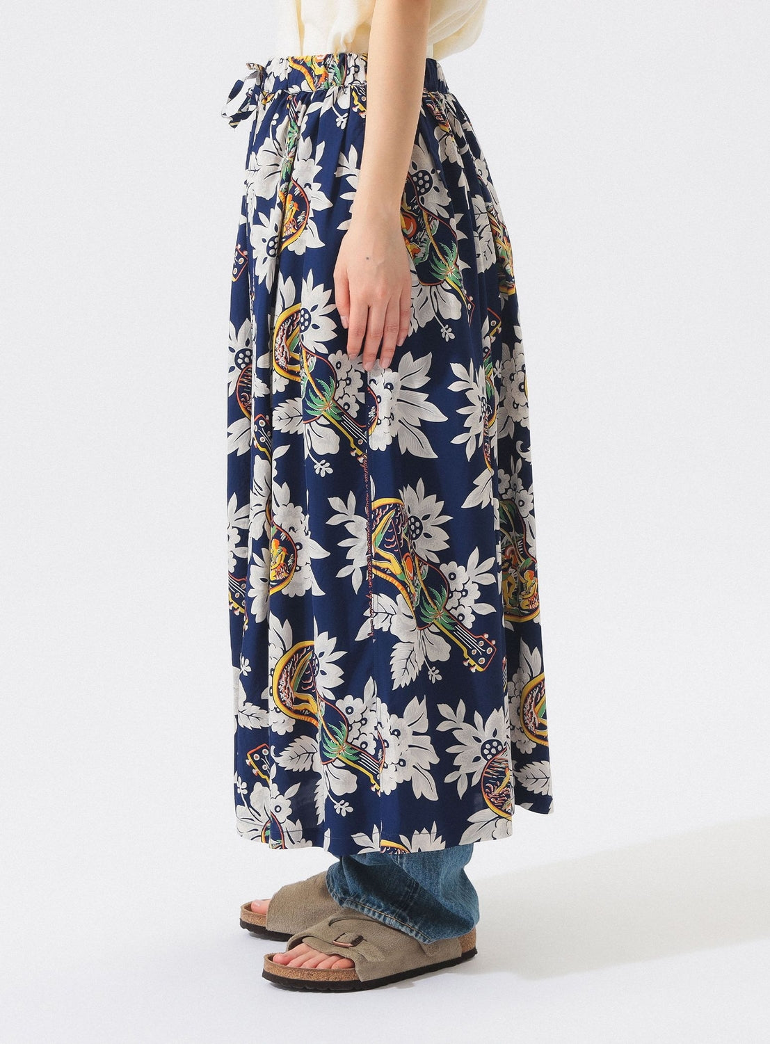 A person stands sideways, showing only their lower half against a white background. They wear the [Exclusive] SUN SURF x BEAMS BOY Aloha Gathered Long Skirt—vintage-inspired prints—over blue jeans, paired with beige suede sandals.