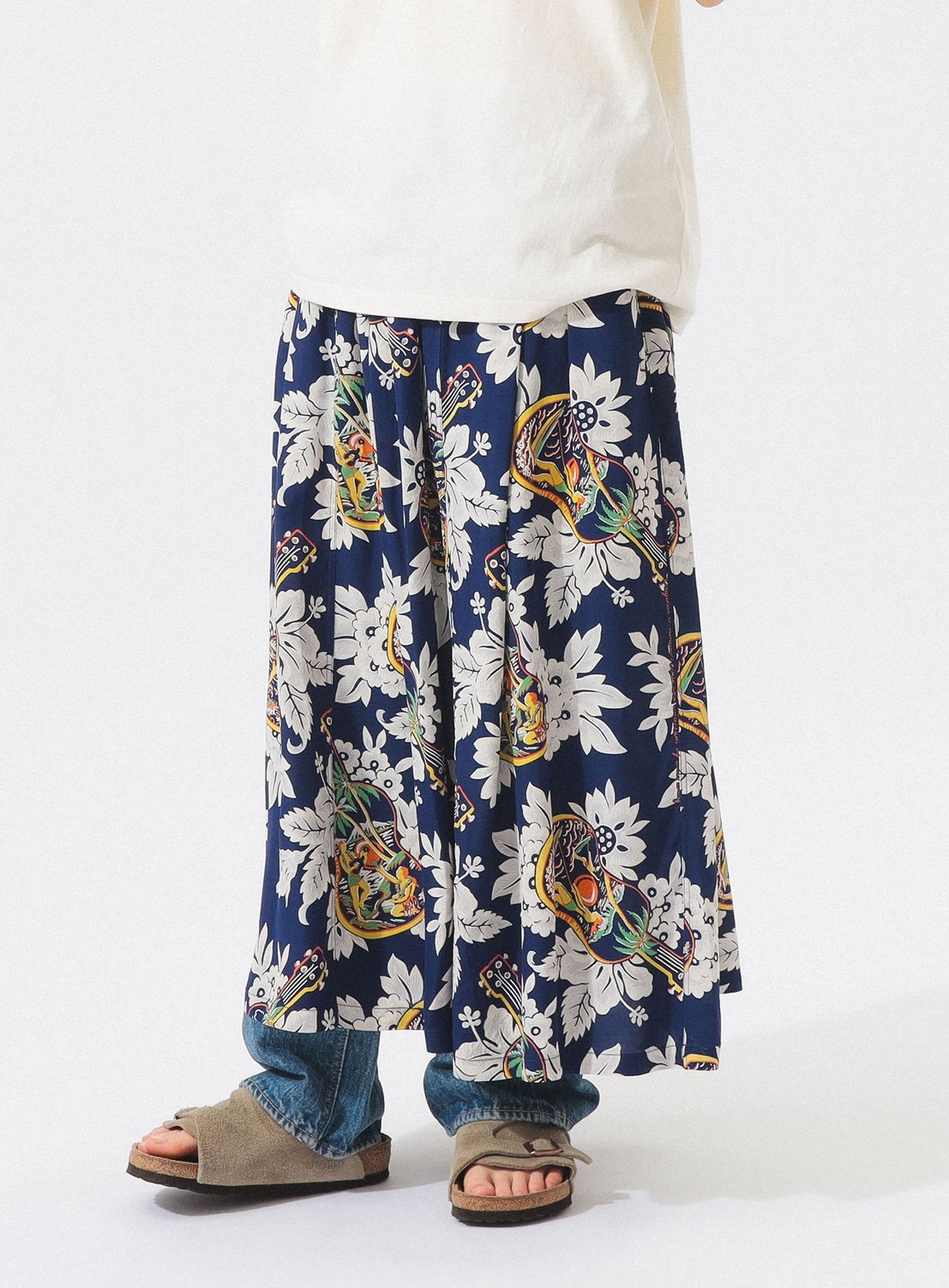 Someone wearing a white top, blue jeans, brown open-toe sandals, and the [Exclusive] SUN SURF x BEAMS BOY Aloha Gathered Long Skirt by SUN SURF with vintage-inspired Hawaiian prints, bold white florals, and colorful birds against a plain background.