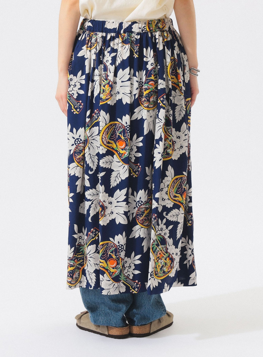 A person wears the [Exclusive] SUN SURF x BEAMS BOY Aloha Gathered Long Skirt by SUN SURF, showcasing a vintage-inspired paisley and white floral print over blue jeans with brown sandals. Only the lower half appears against a white background.
