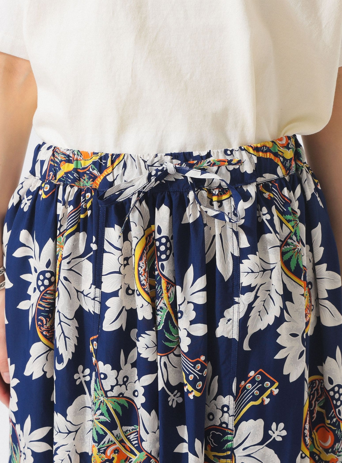 A close-up of a person wearing a white top and the [Exclusive] SUN SURF x BEAMS BOY Aloha Gathered Long Skirt by SUN SURF, featuring vintage-style Hawaiian floral and leaf prints with a tied waistband.