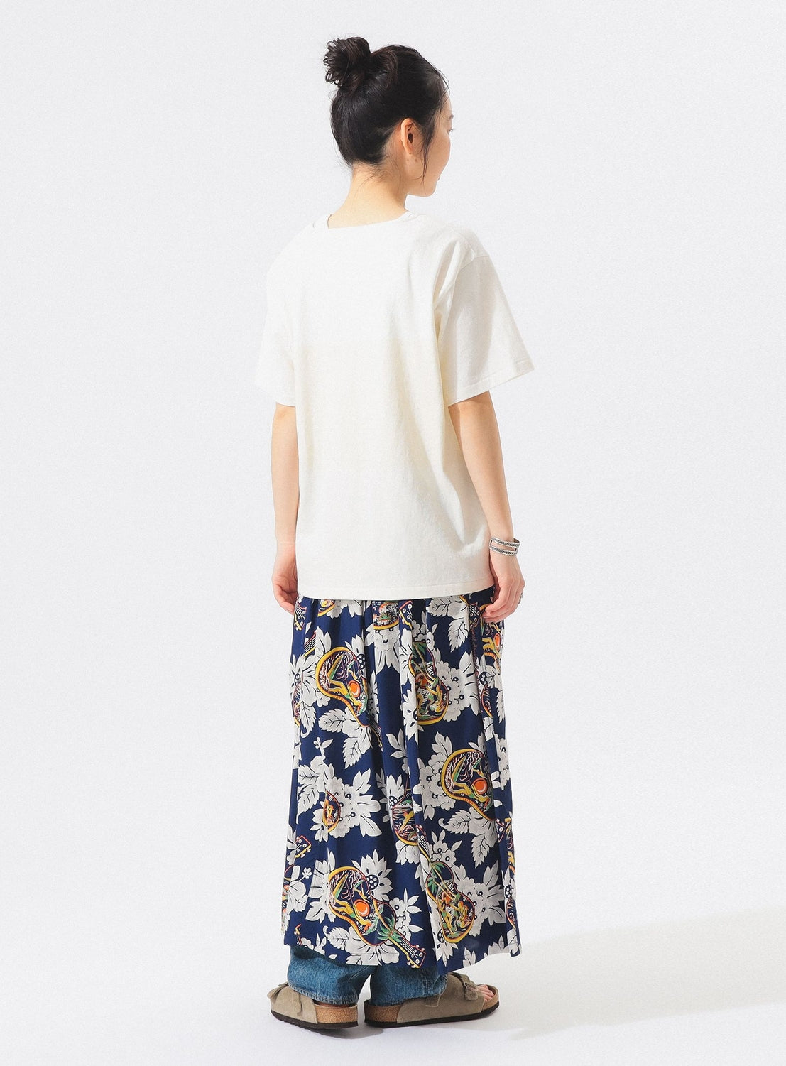 A person stands facing away in a loose white T-shirt, the [Exclusive] SUN SURF x BEAMS BOY Aloha Gathered Long Skirt by SUN SURF over blue jeans, beige sandals, hair in a bun, against a plain white background.