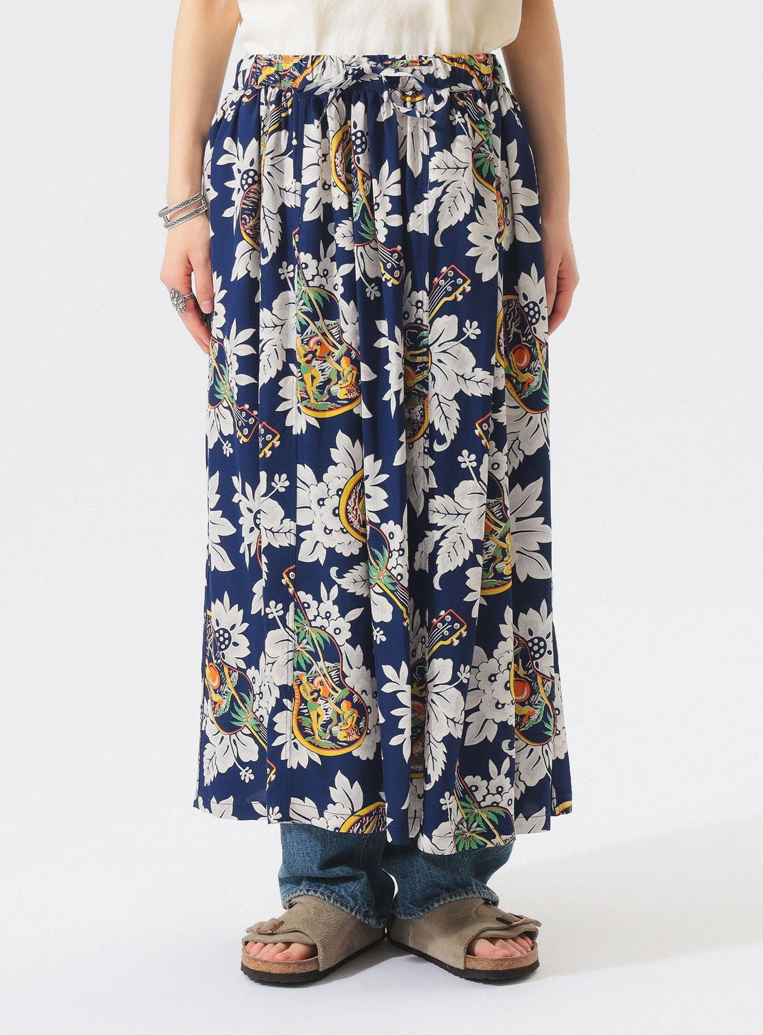 A person wears the [Exclusive] SUN SURF x BEAMS BOY Aloha Gathered Long Skirt with vintage Hawaiian prints, styled with blue jeans, taupe sandals, and bracelets. The photo is cropped at the shoulders to showcase the lower half. Brand: SUN SURF.