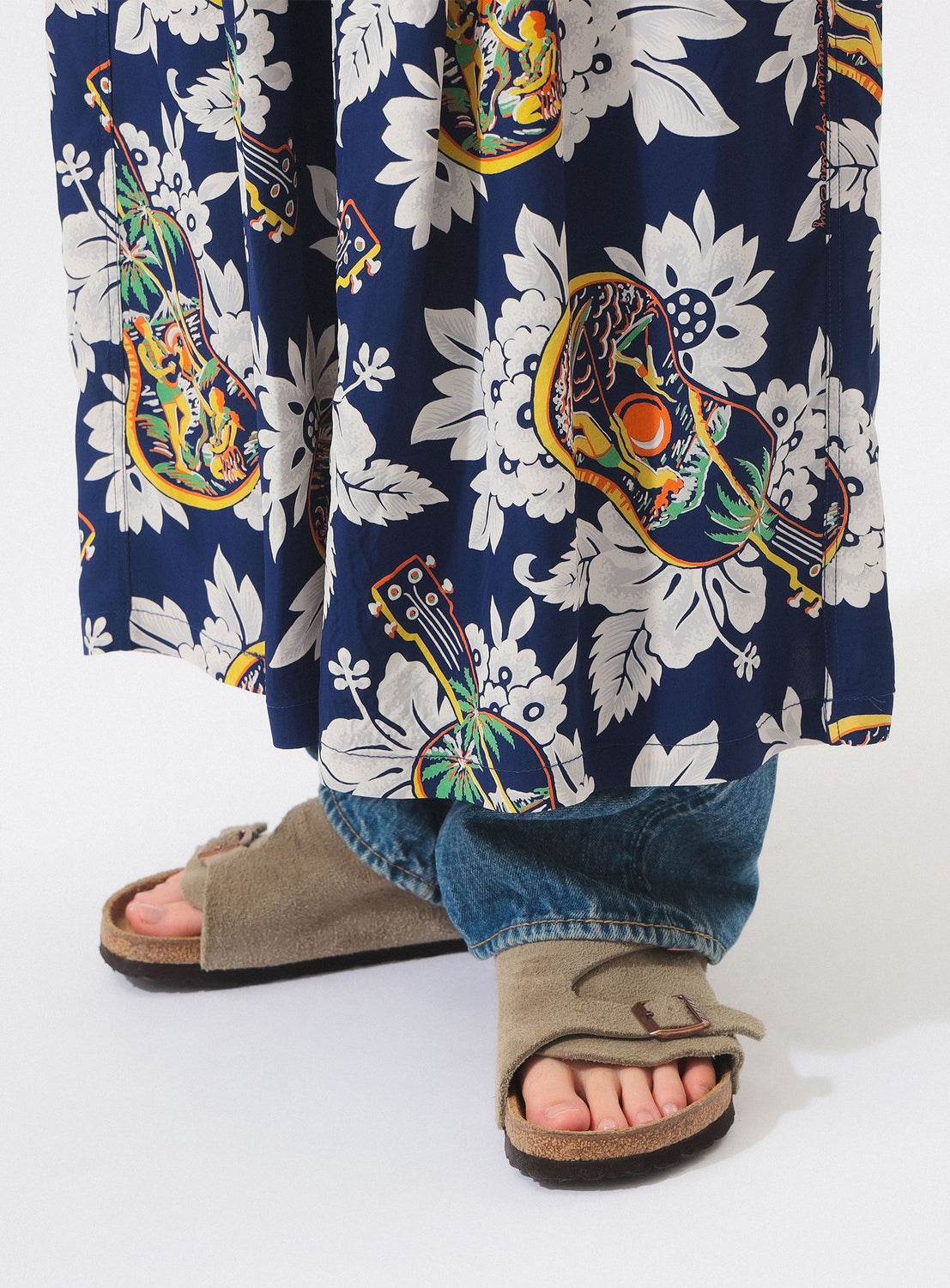 A person wearing the [Exclusive] SUN SURF x BEAMS BOY Aloha Gathered Long Skirt with blue, floral and guitar prints by SUN SURF, paired with loose blue jeans and tan slip-on sandals, stands against a white background.