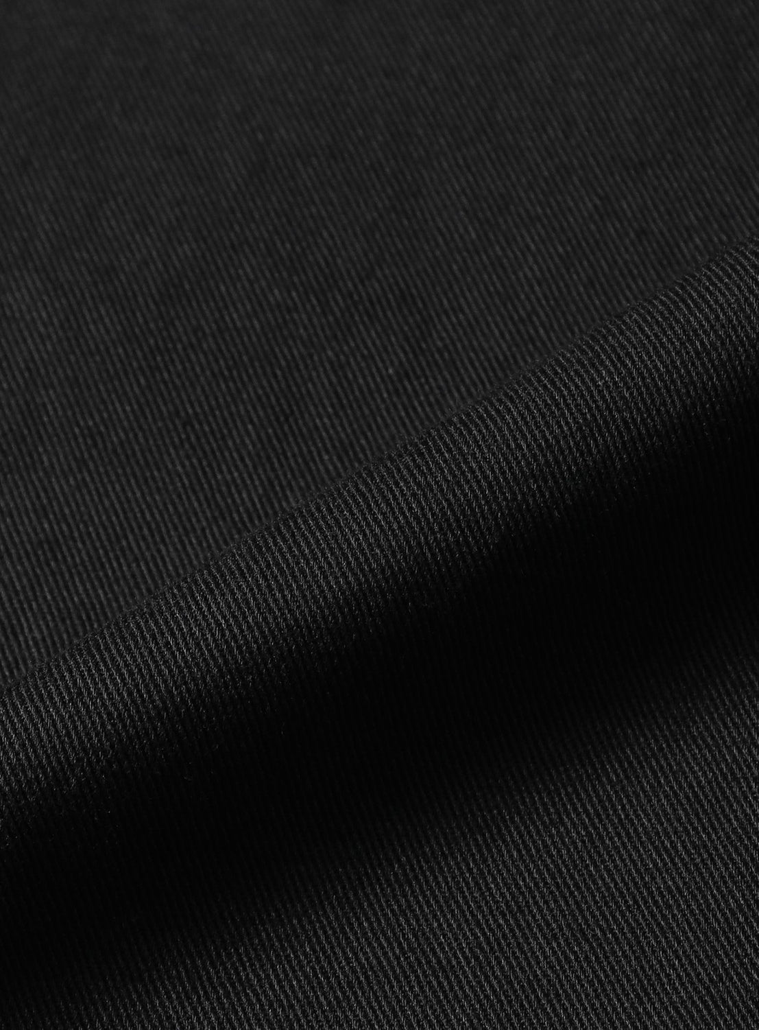 Close-up of black fabric from BEAMS’ Basic Twill Easy Pants, showcasing fine diagonal ridges that indicate a classic twill weave. The material is slightly folded, creating shadows that highlight the texture and stitching details characteristic of relaxed fit designs.