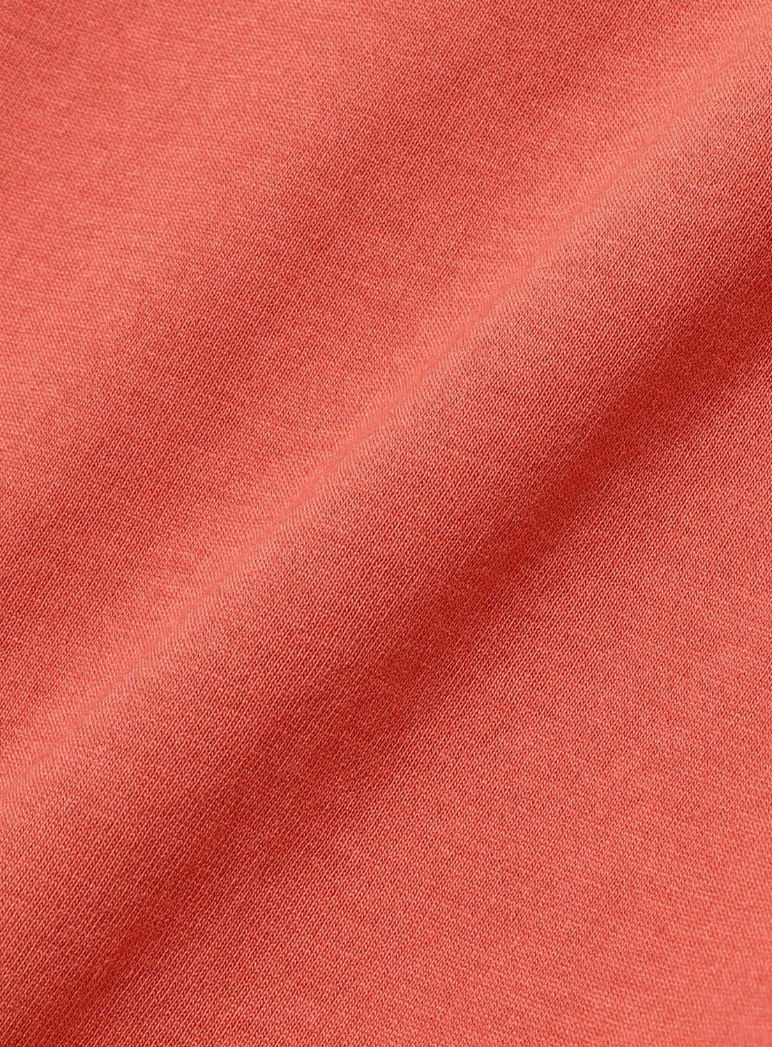 Close-up of a smooth, coral-colored fabric with a subtle diagonal fold highlights the fine woven texture and even color of the BEAMS BOY Two-Tone Vintage Raglan T-shirt, suggesting a soft material ideal for clothing or upholstery and evoking the worn-in look of a classic vintage tee.