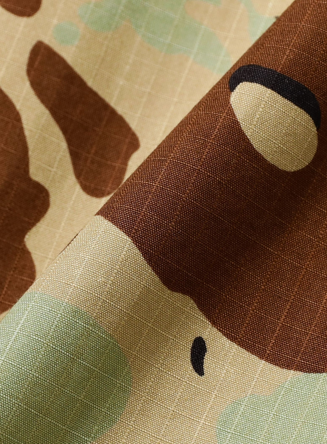 A close-up of NOMARHYTHM TEXTILE’s Camo Flower Open Shirt fabric shows brown, tan, green, and black camouflage with a grid texture. The breathable cotton is softly folded, highlighting its pattern and comfort.