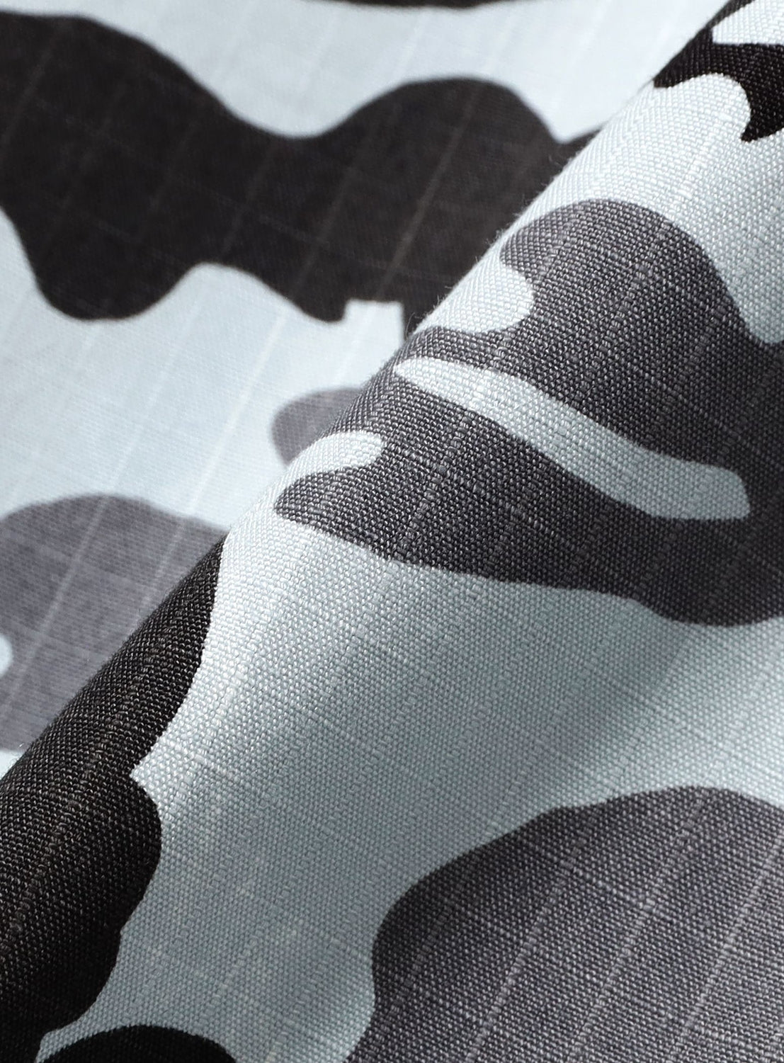 Close-up of light blue, gray, and black camouflage-patterned fabric with a visible grid texture and a soft diagonal fold—highlighting the lightweight cotton featured in the NOMARHYTHM TEXTILE Camo Flower Open Shirt, made in Japan.