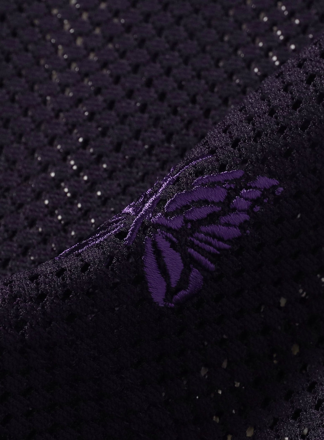 Close-up of the NEEDLES Long Sleeve Geo Raschel Lace Tee in black mesh, showcasing an embroidered purple butterfly motif. Light filters through the geometric lace fabric, making this top ideal for sheer layering or for adding a statement detail to a U-neck T-shirt.