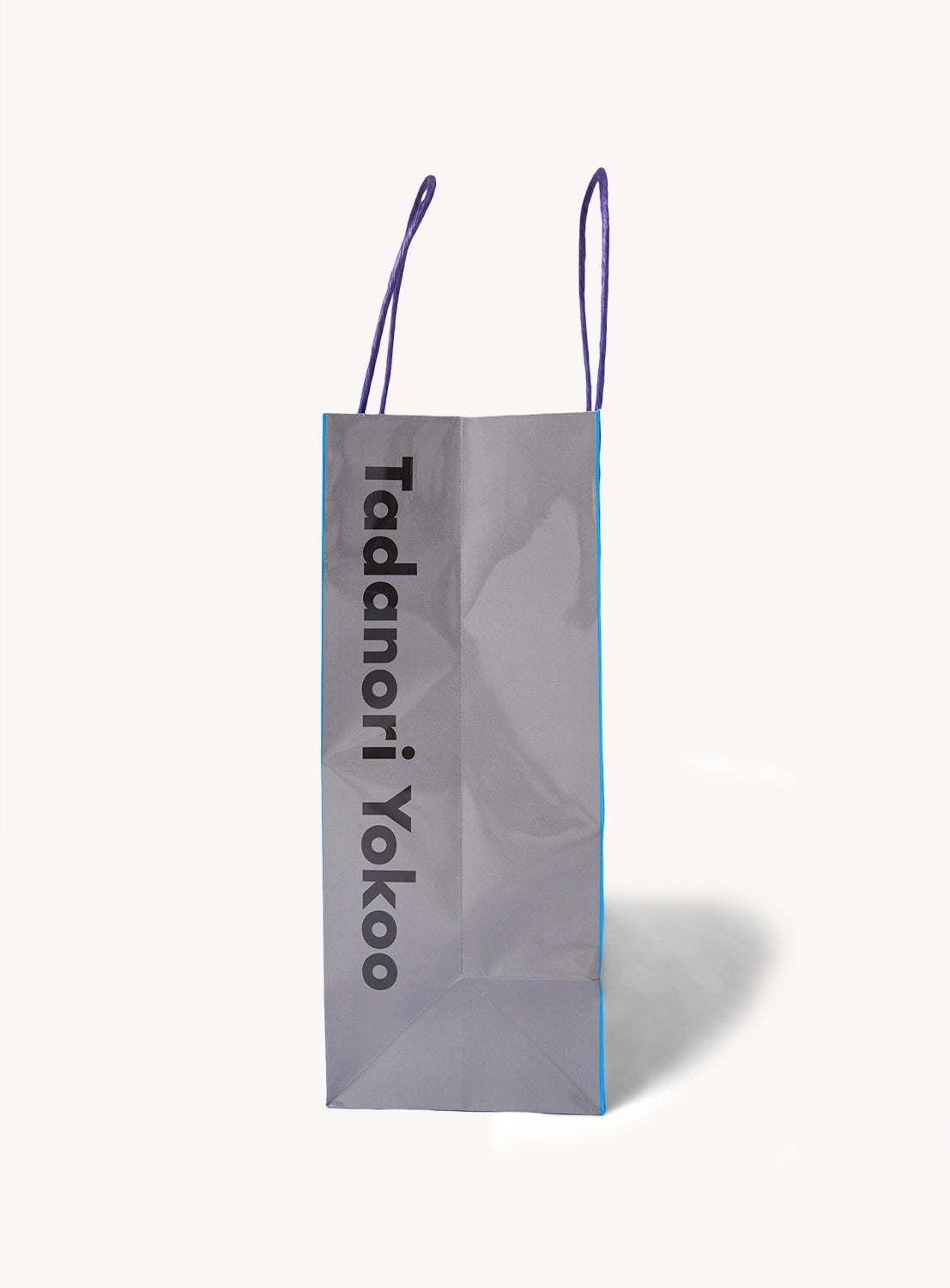 A gray Tadanori Yokoo Shopper Bag from TOKYO CULTUART by BEAMS stands upright, featuring purple handles and “Tadanori Yokoo” printed vertically in bold black letters on the front. The background is plain white.