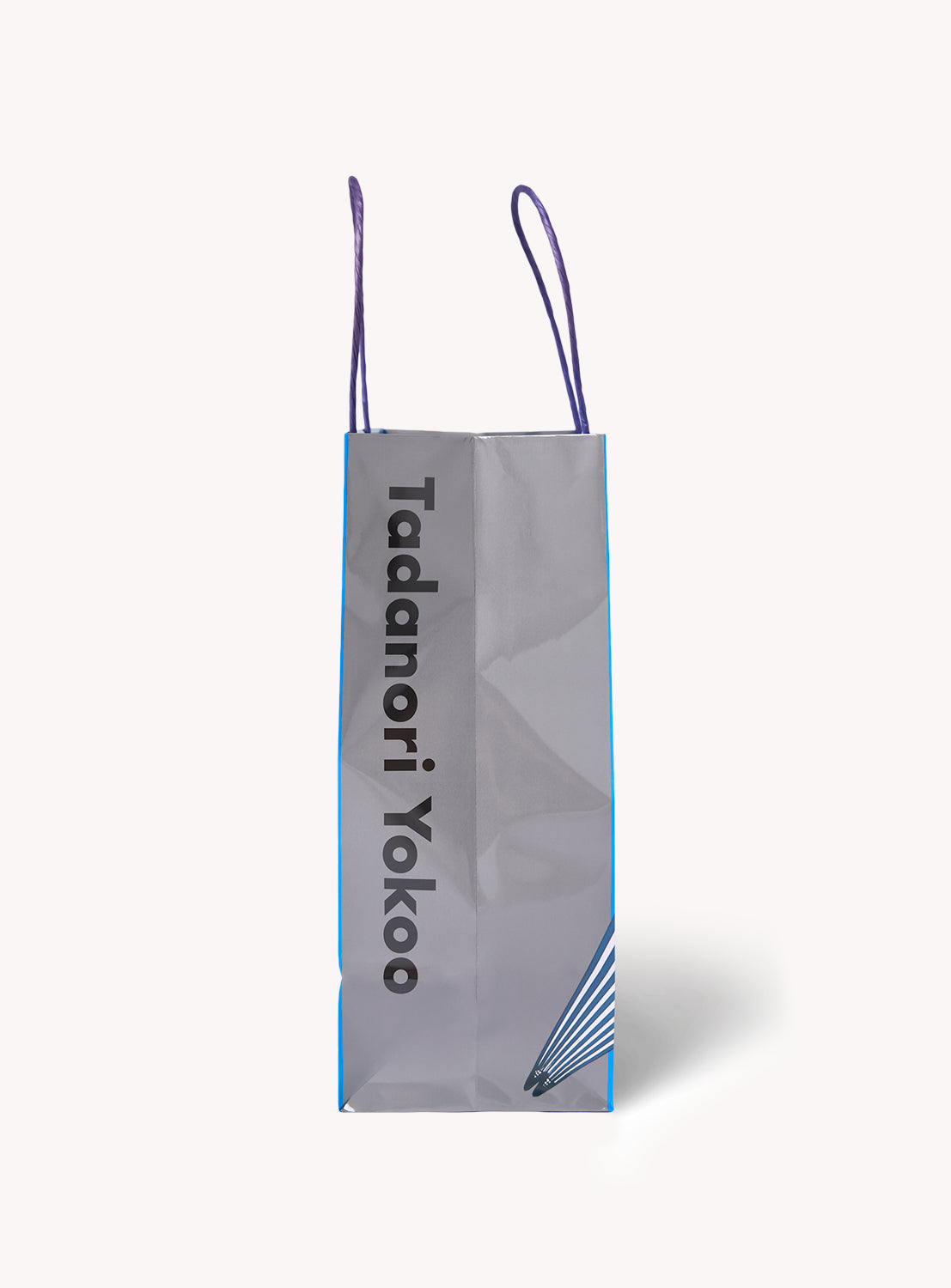 A tall, gray Tadanori Yokoo Shopper Bag by TOKYO CULTUART by BEAMS features purple handles and bold black "Tadanori Yokoo" text down the front, with a blue geometric City and Design-inspired motif accenting the bottom right corner.