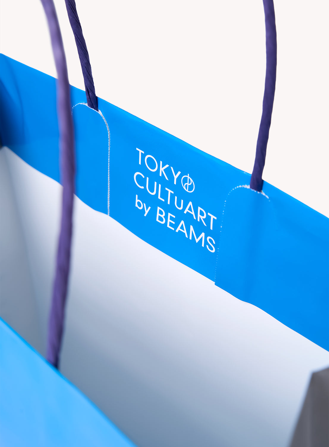 The Tadanori Yokoo Shopper Bag by TOKYO CULTUART by BEAMS is a blue shopper with purple handles, featuring "TOKYO CULTUART by BEAMS" in white text printed inside near the top edge—showcasing city and design motifs inspired by Japanese artist bag aesthetics.