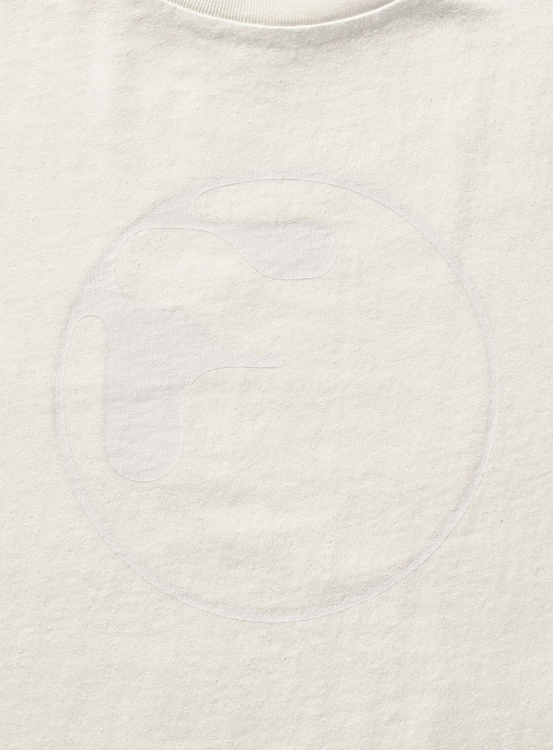 A close-up of the off-white Symbol Logo Tee by F-LAGSTUF-F, featuring a faint, abstract circular logo outlined in white thread near the center—reminiscent of a vintage-inspired graphic highlighted by its unique pigment-dye finish.