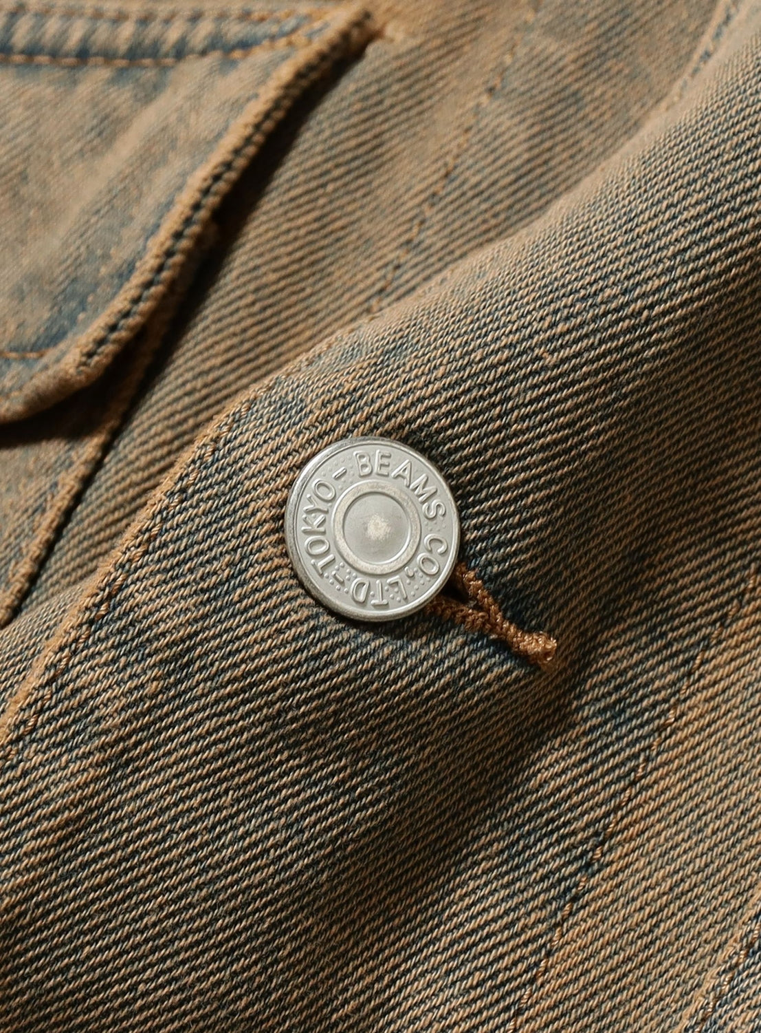 Close-up of the Faded Denim Relaxed Fit Coverall Jacket by BEAMS, highlighting a silver button engraved with BEAMS CO. LTD TOKYO. The lightweight fabric features visible stitching and textured weave, inspired by vintage workwear.