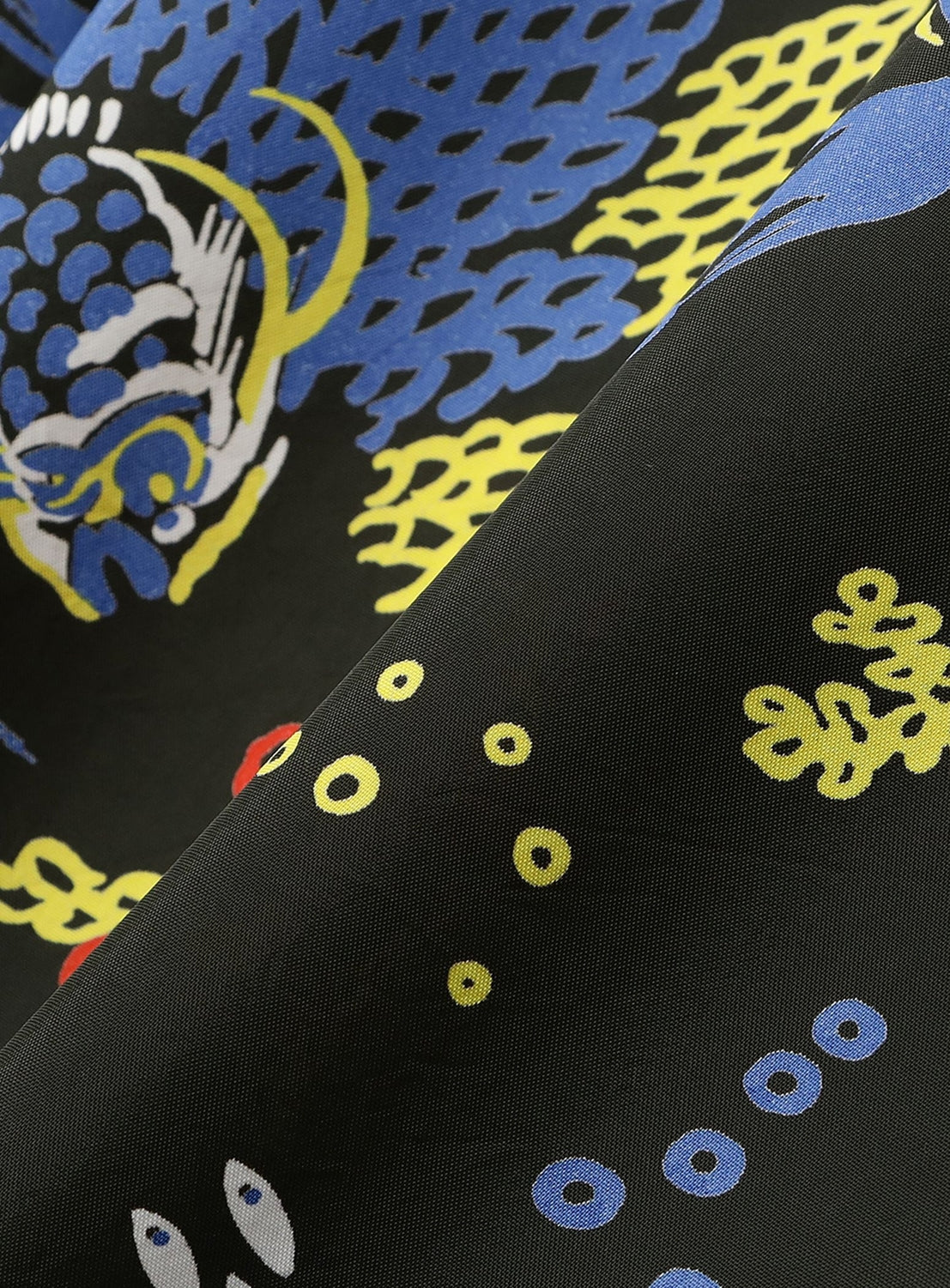 A close-up of black fabric from the SUN SURF Exclusive Patchwork Aloha Shirt 26SS, showing bold, colorful vintage-inspired patterns—abstract blue shapes, yellow dots, and a stylized animal motif on soft material with a slight fold.