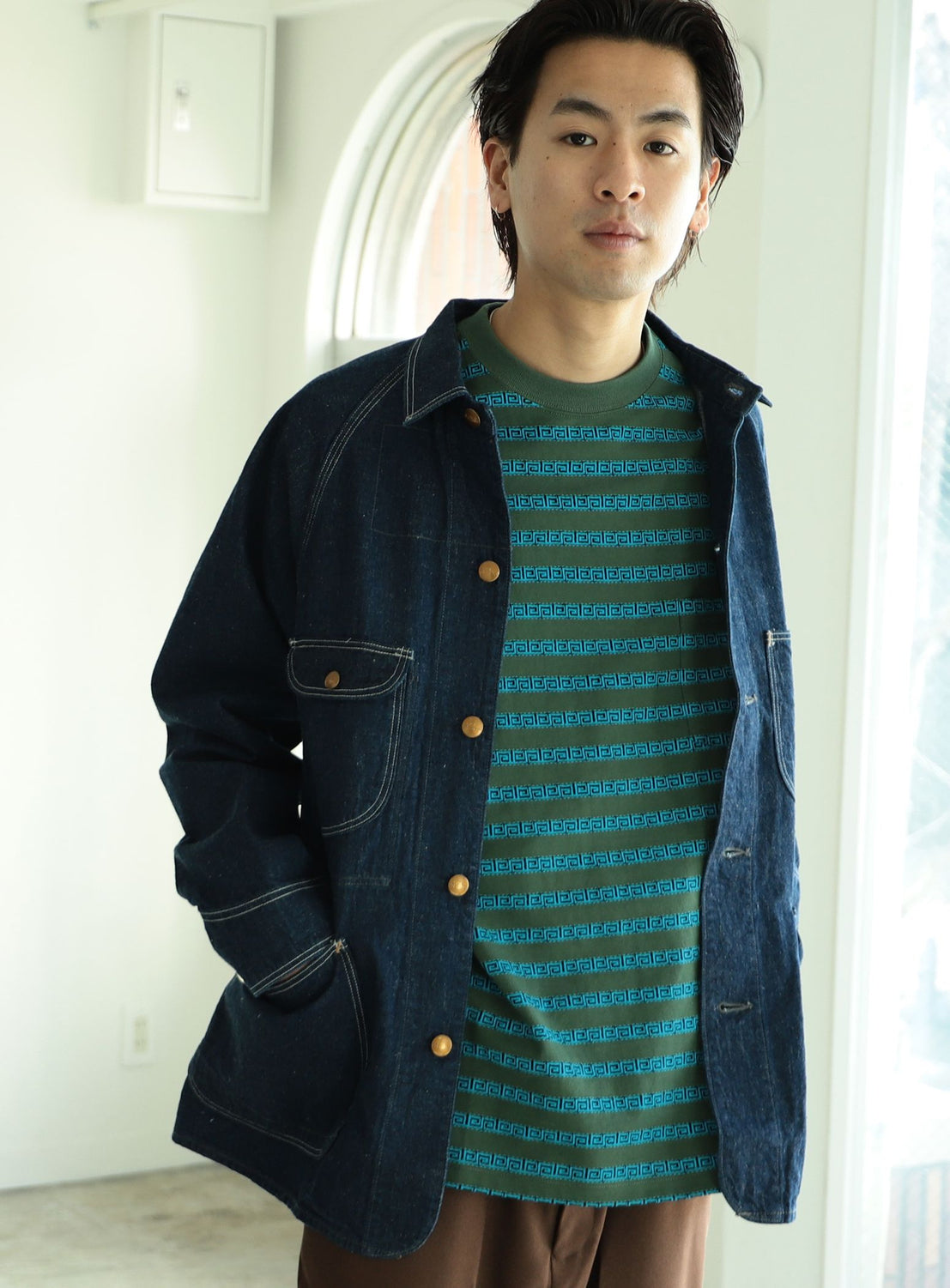 A young man stands indoors with one hand in his pocket, wearing the orSlow 1950 Denim Coverall Jacket—crafted in Japan—layered over a green and blue striped shirt, as sunlight streams through the window behind him.