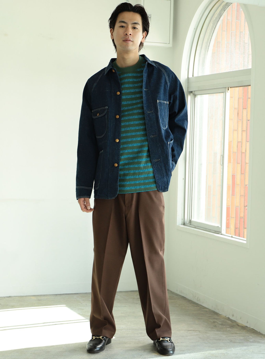 A man stands indoors by a window, wearing the orSlow 1950 Denim Coverall Jacket made in Japan over a green striped shirt, paired with brown trousers and black shoes.