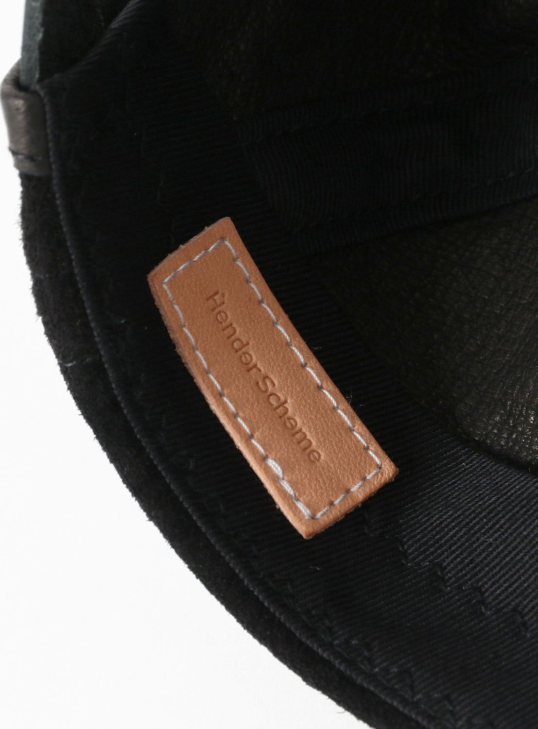 Close-up of Hender Scheme's Pig Jet Cap, a black water-resistant hat with a stitched brown leather tag embossed with the brand’s name. This stylish, durable piece is made in Japan for timeless appeal.