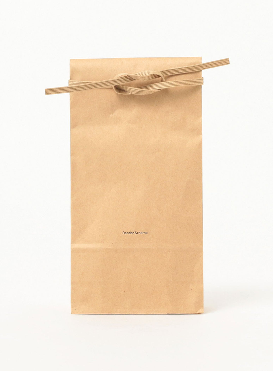 A Fidlock Nylon Belt by Hender Scheme rests upright on a white background, its ribbon-like handle tied in a simple knot. Near the center, small printed text indicates it is made in Japan.