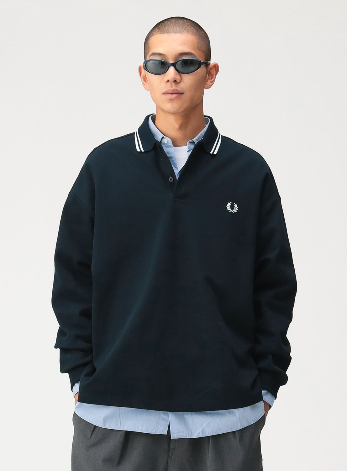 A person wearing an [Exclusive] BEAMS x Fred Perry Pique Long Sleeve Polo Shirt with a twin-tip collar over a light blue collared shirt and dark trousers, finished with dark sunglasses.