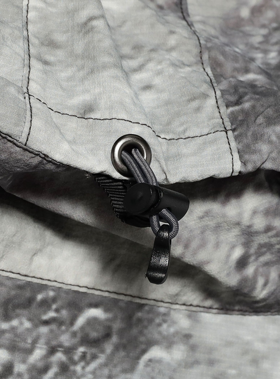 Close-up of a BEAMS Print Zip Hooded Jacket in a grey, mottled-print technical fabric, featuring a metal eyelet, a drawstring cord, and a plastic toggle.