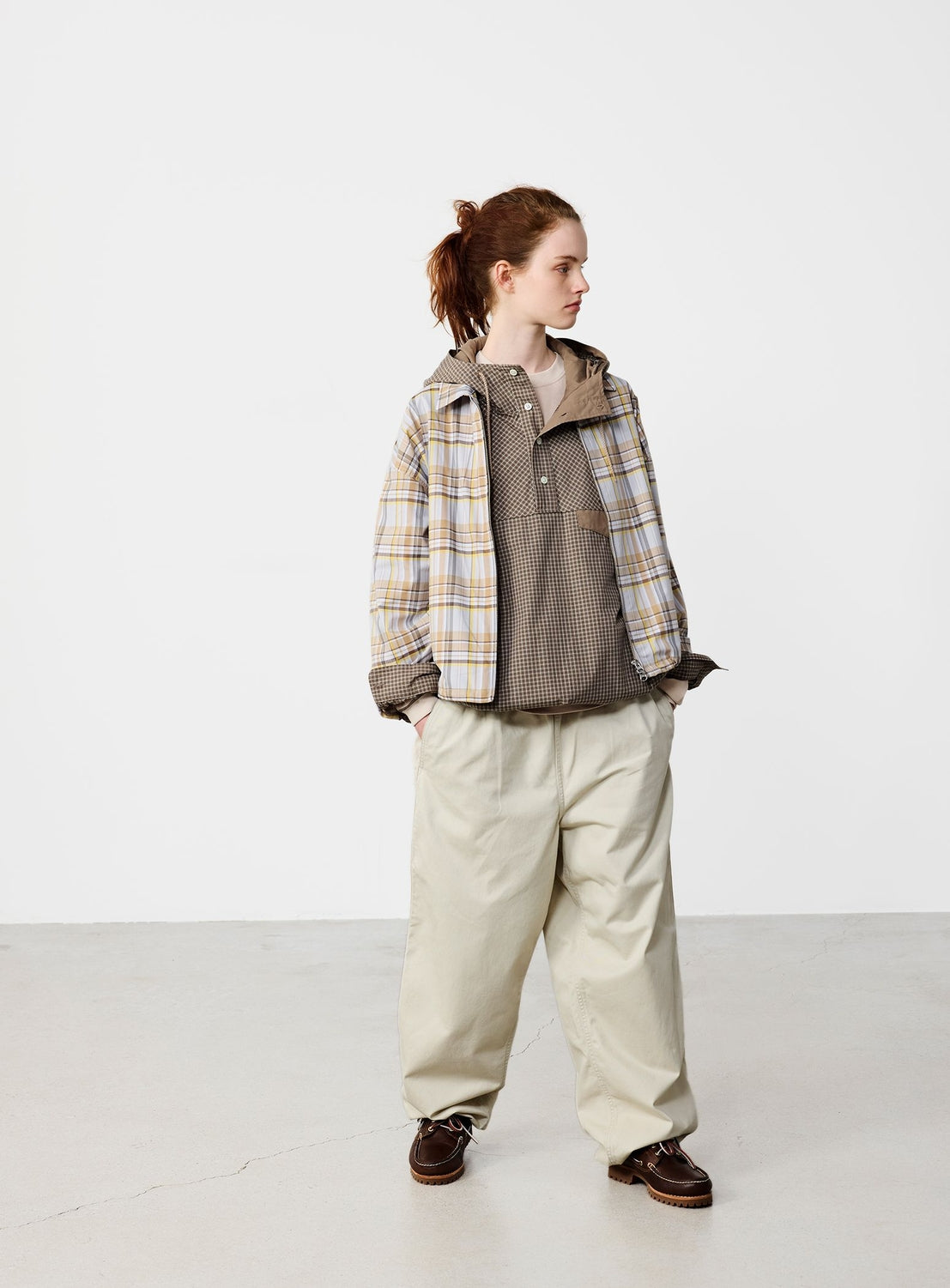 A person stands indoors against a plain white background, hands in pockets and looking to the side, wearing a plaid jacket, checked henley shirt, brown shoes, and BEAMS Basic Twill Easy Pants with an adjustable waistband in a wide relaxed fit.