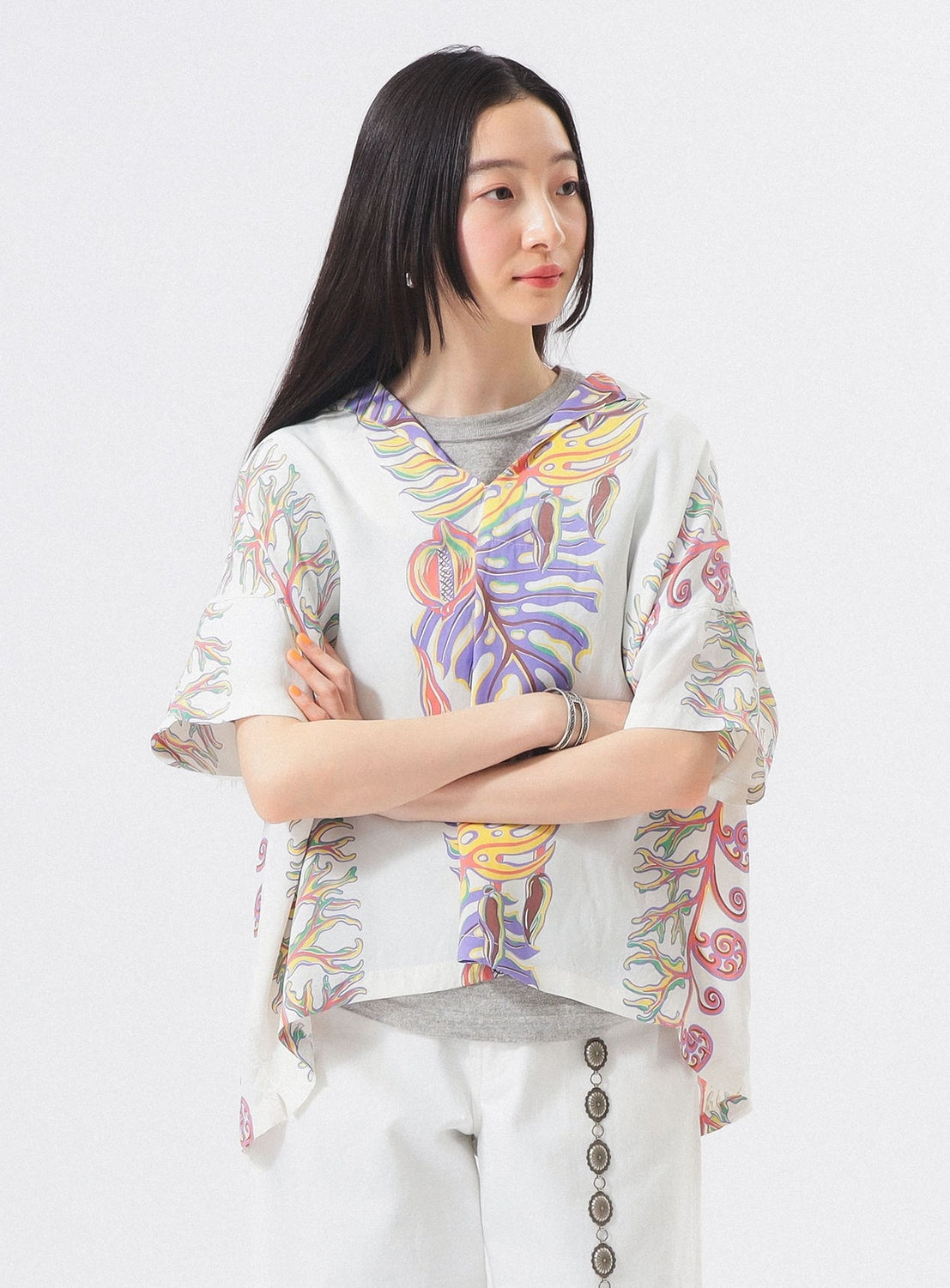 A woman with long dark hair stands arms crossed, wearing the [Exclusive] SUN SURF x BEAMS BOY Sailor Shirt over a gray top and white pants with decorative buttons. She gazes slightly to the side against a plain white background.